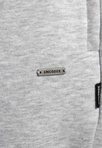 Szary materiał sweatshirtu o gładkiej fakturze. Posiada metalową metkę z napisem "SMILODOX" oraz małą czarną metkę z logo z boku.