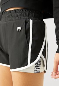 Shorts de sport noirs avec des rayures blanches sur les côtés, une taille élastique et un petit logo sur le devant, superposés à des sous-shorts de couleur claire.