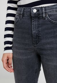 Osoba ubrana w ciemnoszare dżinsy oraz czarną i białą w paski koszulę z długim rękawem, z lewą ręką luźno opartą na boku.