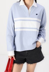 Polo shirt a maniche lunghe azzurro chiaro con strisce bianche, colletto bianco e un piccolo logo a forma di cuore navy. Abbinato a pantaloni shorts neri con orlo sfrangiato.