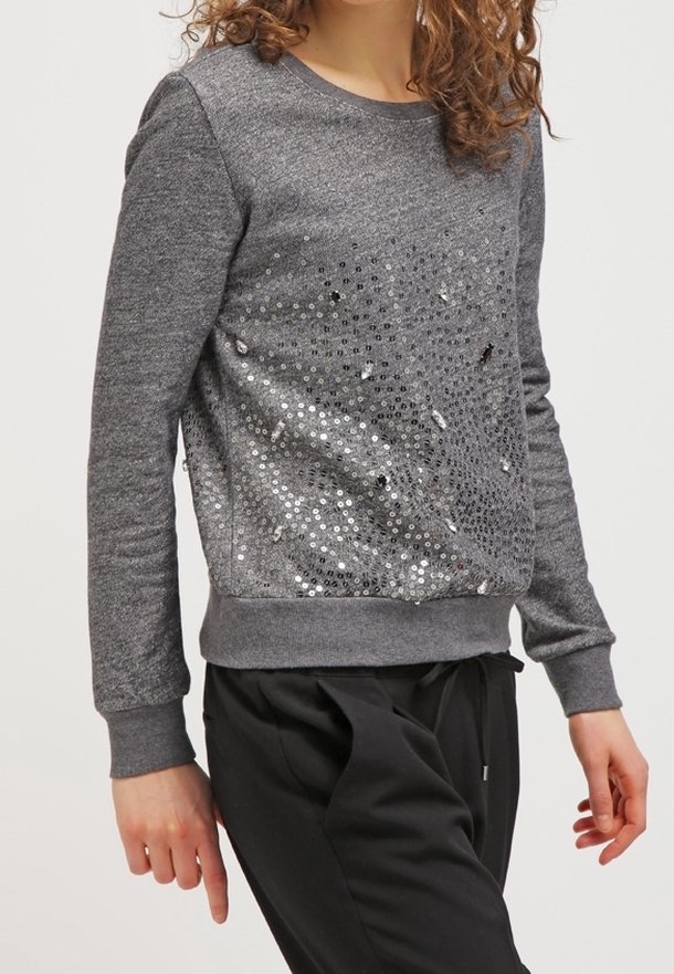 Femme portant un sweat-shirt gris à manches longues avec des sequins argentés sur le devant et un pantalon noir sur un fond uni.