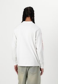 Long-sleeve white cotton t-shirt featuring a crew neckline and printed design along the right sleeve. Loose fit, paired with light blue jeans.