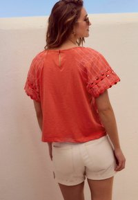 Koraal kleurig top met korte mouwen en geborduurde kanten afwerking, met een sleutelgatdetail op de rug. Gecombineerd met witte opgerolde denim shorts.