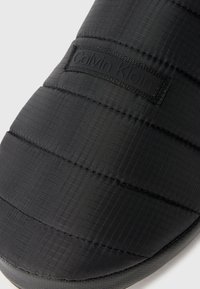 Pantof cu talpă moale, negru, din material textil texturat, cu un model subtil în rețea și un logo Calvin Klein embosat, tot negru.
