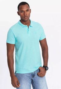 Světle modrá polo košile s krátkým rukávem vyrobená z prodyšného materiálu, s klasickým límcem a logem na hrudi. V kombinaci s denimovými džíny.