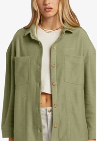 Ljusgrön, texturerad skjortjacka med två bröstfickor och beige knappar, som har en avslappnad passform och krage.