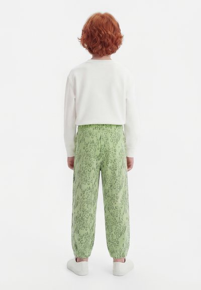 Dítě s kudrnatými zrzavými vlasy stojí zády, oblečené v bílém tričku s dlouhým rukávem, zelených kalhotách s potiskem a bílých botách na bílém pozadí.