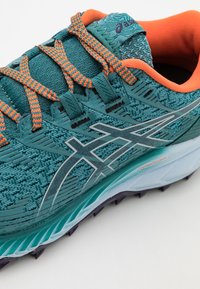 Chaussure de sport teal avec des accents orange, tige en mesh texturé, logo réfléchissant argent, et semelle légère avec un motif en bleu et violet.