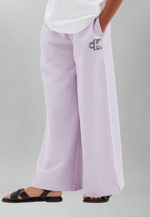 Persona que lleva pantalones lavanda de pierna ancha con un pequeño logo "cK" en el bolsillo, combinados con sandalias negras y una camiseta blanca.