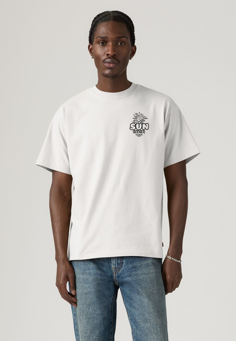 Mand med flettet hår iført hvid T-shirt med "Sun Seeker" solgrafik på brystet og blå jeans mod en ensfarvet baggrund.
