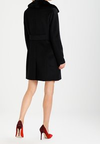 Zwarte wollen jas met een bontkraag, een ceintuurlengte en een rechte snit. Gekozen in combinatie met rode en bordeauxrode hoge hakken.