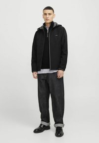 Giacca nera impermeabile con cappuccio grigio, zip frontale e due tasche. Indossata sopra un maglione nero e una camicia a righe, abbinata a jeans scuri e scarpe.