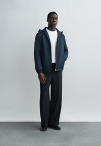 Uomo in piedi davanti a uno sfondo semplice, indossa una giacca con cappuccio blu navy sopra una camicia bianca, pantaloni neri a gamba larga e scarpe nere.