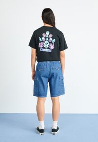 Tricou negru cu un grafic colorat și textul "Converse" pe spate, asortat cu șorturi cargo din denim albastru și adidași negri, șosete albe vizibile.
