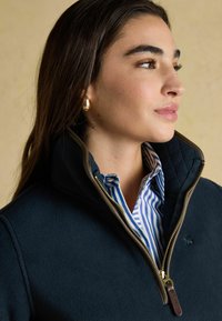 Mladá žena v námornícky modrej fleecovej bunde s polovičným zipsom, oblečená v modro-bielom prúžkovanom košeľovom tričku a zlatých kruhových náušniciach, pozerá doprava.