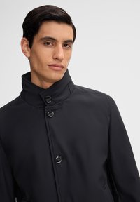 Zwarte waterdichtejas met een hoge kraag, voorzien van een sluiting met knopen aan de voorkant en zijzakken. Glad oppervlak met subtiele stikdetails.