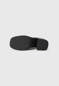 Suola nera della scarpa con texture scanalata sul tallone e sulla parte anteriore, tacco basso e forma rettangolare arrotondata su sfondo chiaro e senza fronzoli.