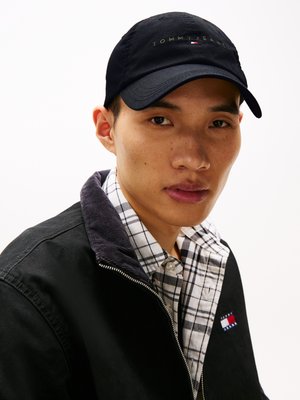 Casquette noire avec "TOMMY JEANS" brodé en or, portée sur une veste noire à col et une chemise à carreaux, montrant un gros plan du visage.