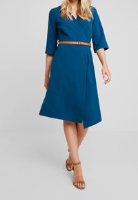 Kvinna som bär en knälång teal wrap-klänning med trekvartslånga ärmar, tanbälte och tanöppna klackar med ankelsnörning, stående framför en enfärgad bakgrund.