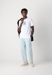 Young man wearing white Calvin Klein t-shirt, light blue jeans, beige sneakers, holding a black jacket over his shoulder, standing against white background.