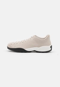 Chaussure de sport beige avec une tige en mesh, une surface texturée, un bout arrondi et une semelle en caoutchouc noire. Présente un accent blanc sur le talon.