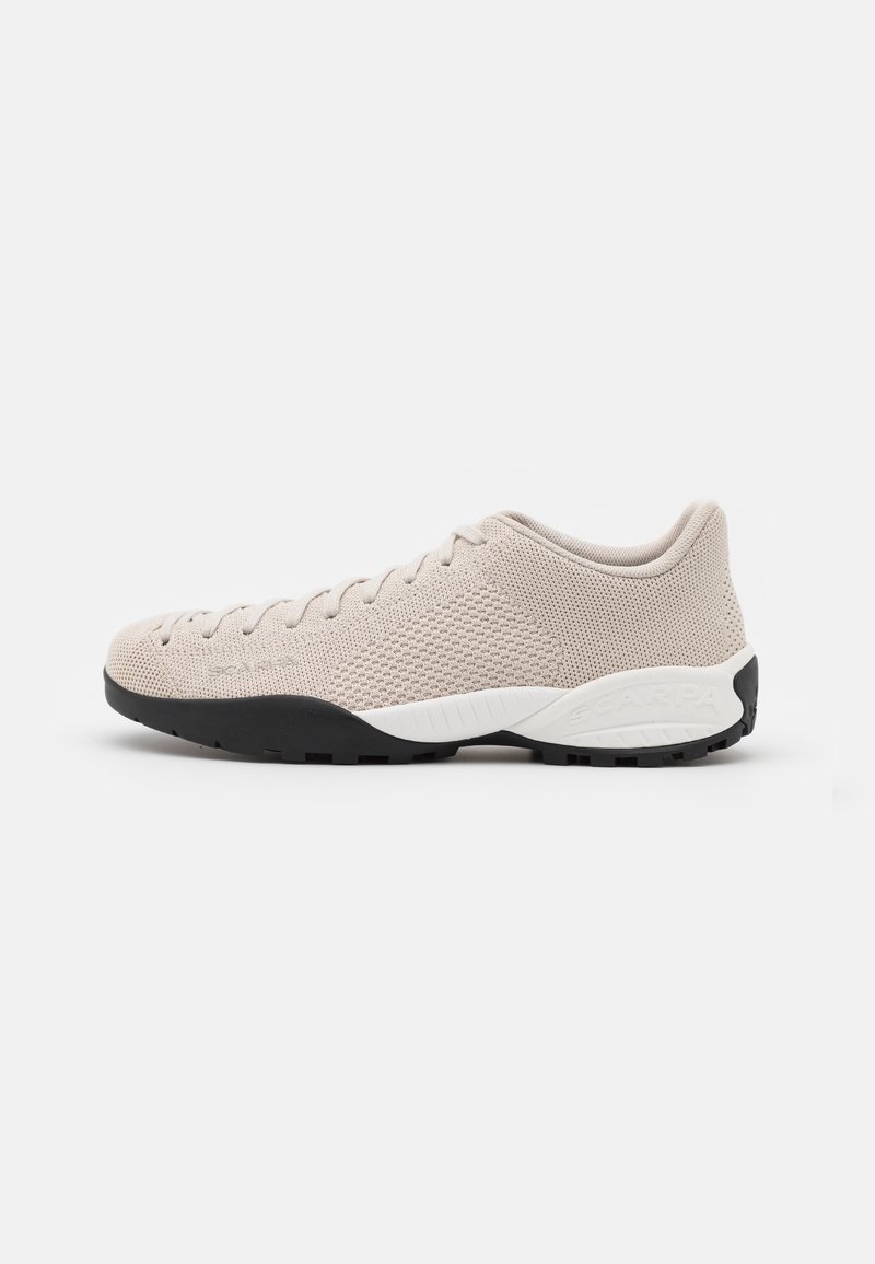 Chaussure de sport beige avec une tige en mesh, une surface texturée, un bout arrondi et une semelle en caoutchouc noire. Présente un accent blanc sur le talon.