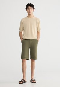 Maglione beige a maniche corte in maglia abbinato a pantaloni corti verdi oliva fino al ginocchio e sandali marroni scuri con una fascia larga.