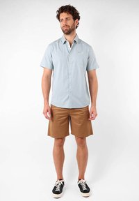 Chemise légère à manches courtes de couleur bleu clair à boutons, associée à un short beige. Des baskets noires avec des accents blancs complètent la tenue.
