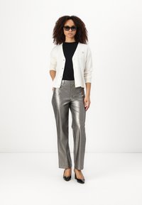 Gilet blanc, t-shirt noir, pantalon large argenté brillant, chaussures à talons noirs, et lunettes de soleil sombres. Coupe décontractée avec des détails côtelés.