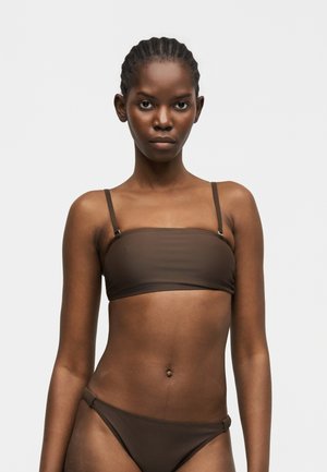 Femeie cu părul împletit, purtând un top bandeau bikini maro închis și slip cu același model, stând în fața unui fundal simplu, deschis la culoare.