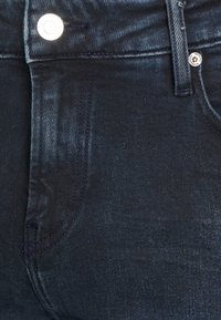 Detailní záběr na tmavě modré džínové kalhoty s viditelnou přední kapsou, poutkem na opasek a stříbrným kovovým knoflíkem.