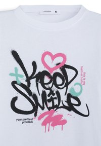 T-shirt bianco in cotone con grafiche testuali colorate in stile spray: "Keep Smile" in nero, con accenti rosa e teal. Etichetta della taglia visibile.