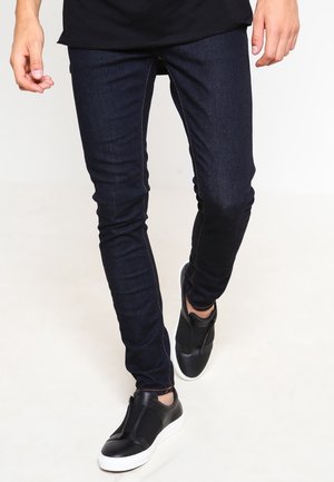 Persona che indossa jeans aderenti blu scuro e sneaker slip-on nere che cammina su uno sfondo chiaro.