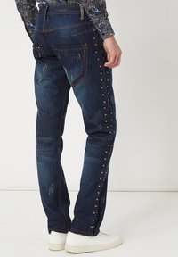 Jeans de mezclilla azul oscuro con detalles desgastados, tachuelas en los laterales y dos bolsillos traseros. Llevados con zapatillas blancas y una camisa estampada.