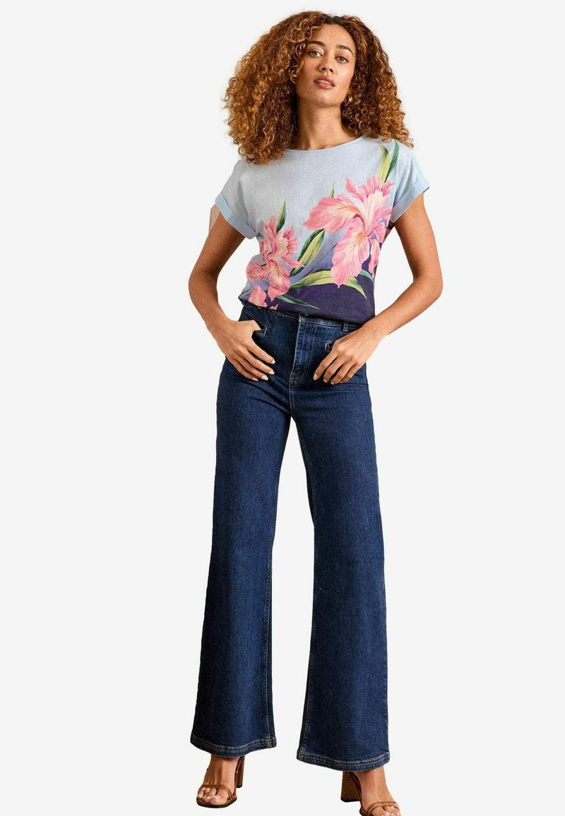 Gėlių raštais margintas šviesiai mėlynas viršus su trumpomis sukritusiomis rankovėmis, derinamas su tamsiai mėlynomis plačiomis kelnėmis. Modelis stovi platforminėse šlepetėse.