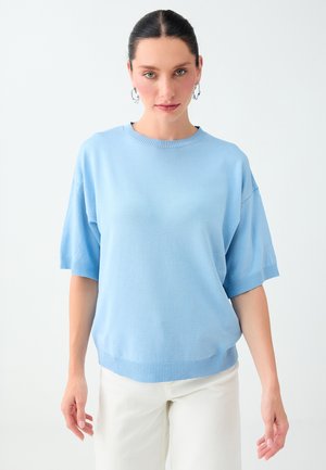 Vrouw met donker haar in een blauw kortemouwen trui en witte broek, staand tegen een effen lichte achtergrond.