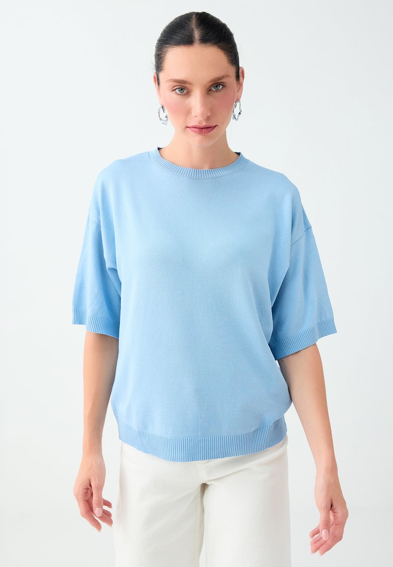 Femme aux cheveux foncés portant un pull bleu à manches courtes et un pantalon blanc, debout devant un fond clair uni.