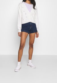 Cárdigan blanco de punto con bordado floral, combinado con pantalones cortos de mezclilla oscuros y zapatillas altas blancas. Diseño de cuello en V y botones visibles.