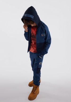 Ensemble de sweat à capuche et de pantalon bleu marine avec des accents bleu foncé, comportant un logo sur la capuche et un patch sur le vêtement ; assorti à des bottes beiges.