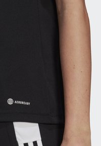 Närbild av en persons arm bredvid en svart tröja med "AEROREADY"-logotyp och svartvita randiga sportshorts.