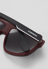 Punased prillid tumedate glasuuridega, millel on must kitsas käsi, kaunistatud hõbedaste detailidega ja valge "CARRERA" logo.