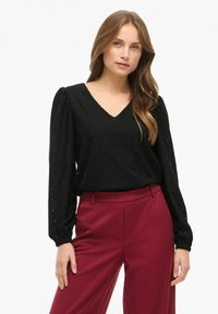 Top nero a V con motivo a losanghe e maniche lunghe a sbuffo, abbinato a pantaloni bordeaux a vita alta in taglio dritto.