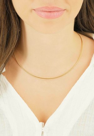 Collana choker in metallo dorato, superficie liscia, design circolare, si adatta strettamente attorno al collo. Indossata con una blusa bianca texturizzata.
