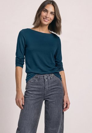 Femme aux cheveux bruns mi-longs portant un t-shirt à manches longues bleu sarcelle foncé et un jean gris taille haute, souriant à la caméra.