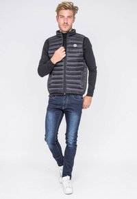 Gilet noir en doudoune avec des coutures horizontales, fermeture zippée à l'avant et un patch logo rond. Assorti à un jean bleu slim et des baskets blanches.