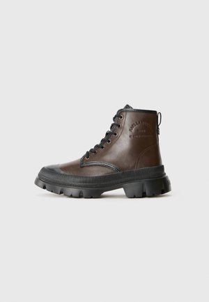 Brown leather ankle boots with black rubber outsole, lace-up design, textured accents, and embossed branding on the side.