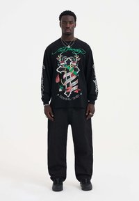 Schwarzes Langarmshirt mit buntem Grafikdruck und Motiven, kombiniert mit lockeren grauen Hosen und schwarzen Sneakers.