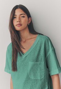 Zelené veľké tričko s výstrihom do V a zavinutými rukávmi. Má texturovanú látku a náprsné vrecko na ľavej strane.