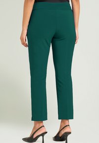 Groene, nauwsluitende broek met een gladde textuur, voorzien van een tailleband op mid-rise hoogte en smalle openingen bij de enkels, gecombineerd met zwarte schoenen met hoge hakken.