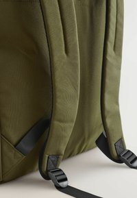 Nahrbtnik iz tkanine olivne zelene barve z črnimi nastavljivimi naramnicami, ojačanimi šivi in kovinskimi dodatki. Ima teksturirano površino.